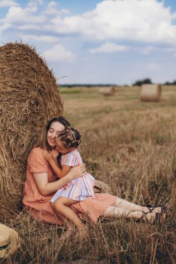 Anne ve kızı hasat edilen buğday tarlasında saman balyasının yanında oturuyor. Diğer balyaların arka planına karşı. Kızın, annenin yüzüne bastırılmış.