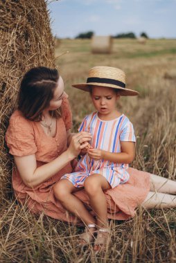 Genç anne ve kız hasat edilen buğday tarlasında saman balyasının yanında oturuyorlar. Diğer balyaların arka planına karşı. Annem kızına saman anahtarı takıyor.