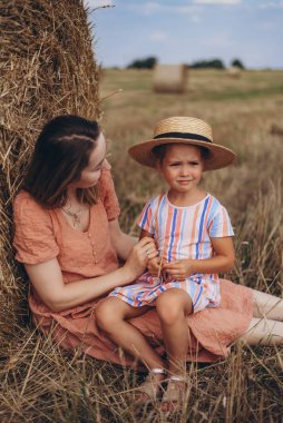 Genç anne ve kız hasat edilen buğday tarlasında saman balyasının yanında oturuyorlar. Diğer balyaların arka planına karşı. Kızım başka tarafa bakıyor.