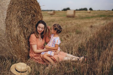 Anne ve kızı hasat edilen buğday tarlasında saman balyasının yanında oturuyorlar. Diğer balyaların arka planına karşı. Kızı annesinin saçına dokunuyor.