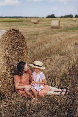 Genç anne ve kız hasat edilen buğday tarlasında saman balyasının yanında oturuyorlar. Diğer balyaların arka planına karşı. Kızım başka tarafa bakıyor.