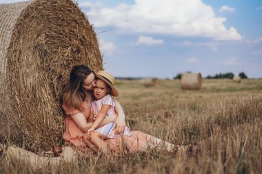 Genç anne ve kız hasat edilen buğday tarlasında bir saman balyasının yanında oturuyorlar. Diğer balyaların arka planına karşı. Anne kızına sıkıca sarılıyor.