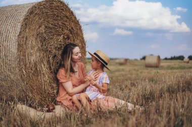 Genç anne ve kız hasat edilen buğday tarlasında saman balyasının yanında oturuyorlar. Anne ve kızı birbirlerine bakıyorlar.
