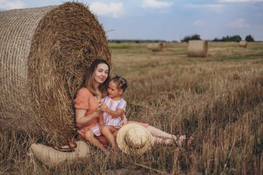 Genç anne ve kız hasat edilen buğday tarlasında saman balyasının yanında oturuyorlar. Kızı annesinin elini tutuyor..