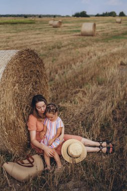 Genç anne ve kız hasat edilen buğday tarlasında saman balyasının yanında oturuyorlar. Yanlarında hasır bir çanta ve bir şapka var.