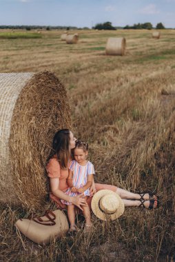 Genç anne ve kız hasat edilen buğday tarlasında saman balyasının yanında oturuyorlar. Yanlarında hasır bir çanta ve bir şapka var.