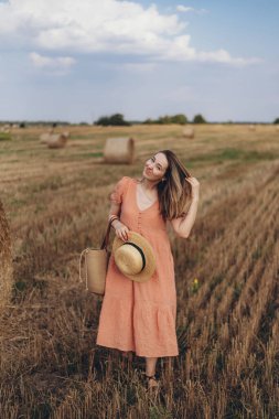 Uzun mercan renkli elbiseli ve uzun saçlı genç bir kız elinde şapkayla tarlada yürüyor.