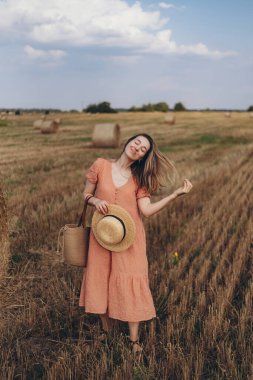 Uzun mercan renkli elbiseli ve uzun saçlı genç bir kız elinde şapkayla bir tarlada yürüyor. Gözlerini kapadı.