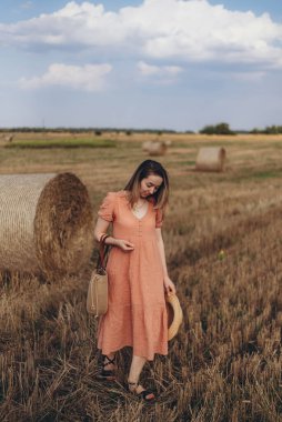 Uzun elbiseli, uzun saçlı, elinde hasır bir çanta tutan genç bir kız. Kız elinde şapkayla sahada dolaşıyor. Arka planda balya samanı