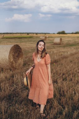 Uzun elbiseli, uzun saçlı, elinde hasır bir çanta tutan genç bir kız. Kız elinde şapkayla sahada dolaşıyor. Arka planda balya samanı