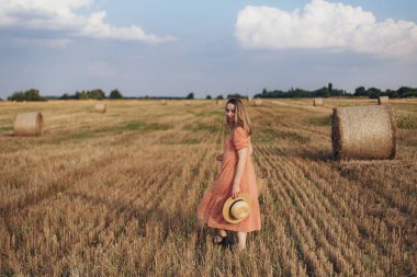 Uzun elbiseli, uzun saçlı, elinde hasır bir çanta tutan genç bir kız. Kız elinde şapkayla sahada dolaşıyor. Arka planda balya samanı