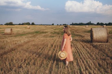 Uzun elbiseli, uzun saçlı, elinde hasır bir çanta tutan genç bir kız. Kız elinde şapkayla sahada dolaşıyor. Arka planda balya samanı