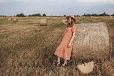 Uzun elbiseli ve uzun saçlı genç bir kız buğday tarlasında bir saman balyasını bulutlandırdı. Hasır şapkalı kız.