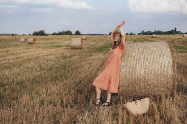 Uzun elbiseli ve uzun saçlı genç bir kız buğday tarlasında bir saman balyasını bulutlandırdı. Hasır şapkalı kız.