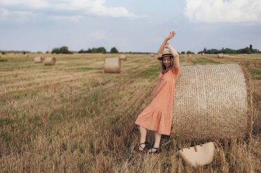 Uzun elbiseli ve uzun saçlı genç bir kız buğday tarlasında bir saman balyasını bulutlandırdı. Hasır şapkalı kız.