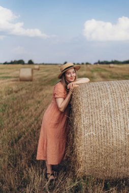 Uzun elbiseli ve uzun saçlı genç bir kız hasat edilen buğday tarlasında bir saman balyasını güzellikle gölgeliyor. Hasır şapkalı bir kız.