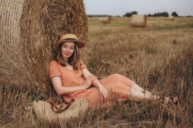 Uzun elbiseli güzel bir kız ve uzun saçlı sevimli bir gülümseme bir saman balyası hakkında bulutlu oturuyor. Kız uzaktan bakar ve hayal kurar.