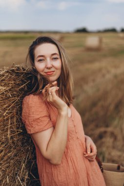 Uzun saçlı, elbise giymiş güzel bir kızın portresi. Kız bir saman balyası hakkında bulutlandı. Arka plan bulanık. Bir bulut arkaplanı karşısında