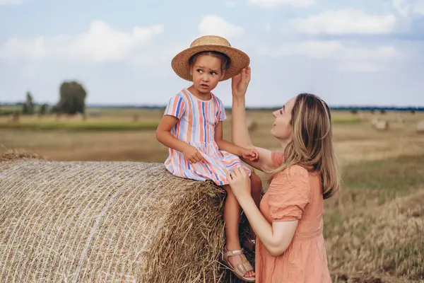 Anne ve küçük kızı mavi gökyüzüne karşı hasat edilmiş buğday tarlasında. Kız bir saman balyasının üzerinde oturuyor. Annem kızına hasır şapka takıyor.