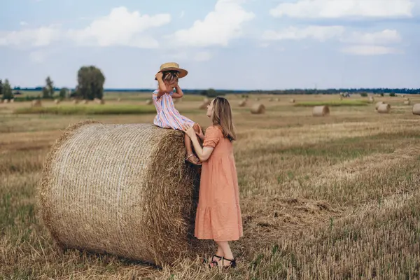 Hasat edilen buğday tarlası. Küçük bir kız bir saman balyasına oturur ve şapkasını kafasına takar. Mercan elbiseli yetişkin bir anne yakınlarda duruyor ve kızına bakıyor.