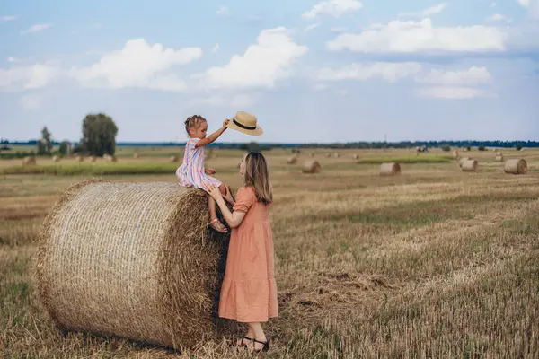 Hasat edilen buğday tarlası. Küçük bir kız bir saman balyasının üzerinde oturuyor. Mercan elbiseli yetişkin bir anne yakınlarda durup kızına bakar. Bir kız annesine şapka takmaya çalışıyor.