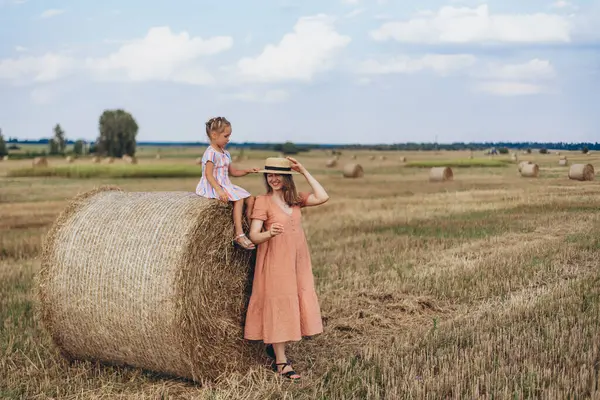 Hasat edilen buğday tarlası. Küçük bir kız bir saman balyasının üzerinde oturuyor. Mercan elbiseli yetişkin bir anne yakınlarda durup kızına bakar. Kız annesinin kafasına bir şapka takıyor. Annem gülümser.