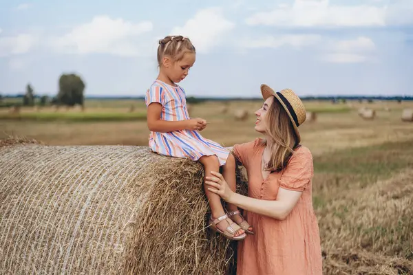 Hasır şapkalı genç bir anne ve hasat edilen buğday tarlasında iletişim kuran genç bir kız, saman balyasında oturan bir kız.