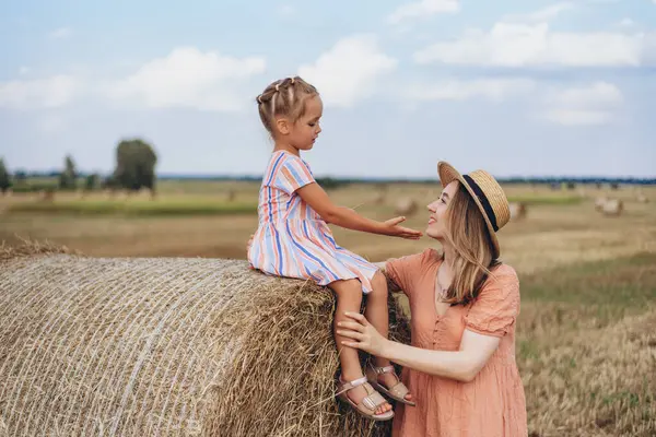 Hasır şapkalı genç bir anne ve hasat edilen buğday tarlasında iletişim kuran genç bir kız, saman balyasında oturan bir kız.