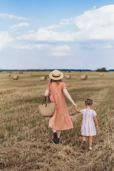 Uzun elbiseli ve hasır şapkalı genç bir anne küçük kızını elinden tutar. Her ikisi de hasat edilen buğday tarlasında yürür. Bir saman balyasının arka planında