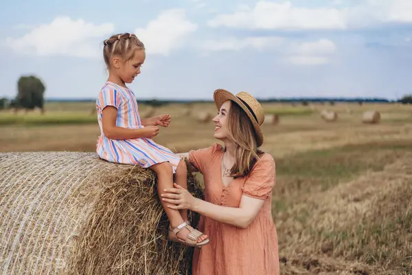 Hasır şapkalı genç bir anne ve hasat edilen buğday tarlasında iletişim kuran genç bir kız, saman balyasında oturan bir kız.