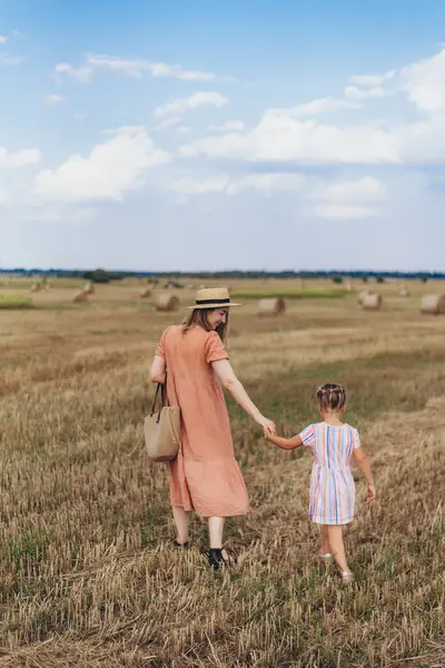 Uzun elbiseli ve hasır şapkalı genç bir anne küçük kızını elinden tutar. Her ikisi de hasat edilen buğday tarlasında yürür. Bir saman balyasının arka planında