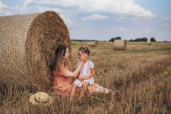 Mother and daughter sitting next to a bale of straw on a harvested wheat field. against the background of other bales. Daughter pressed against the mothers face