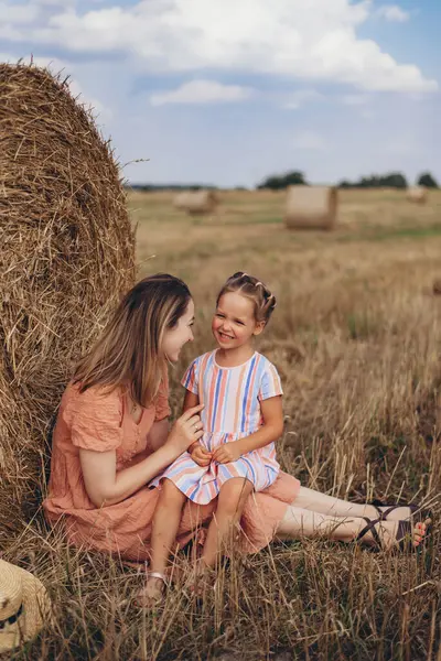 Mother and daughter sitting next to a bale of straw on a harvested wheat field. against the background of other bales. Mother leans back to her daughter. Daughter smiling