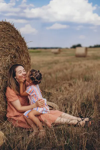 Mother and daughter sitting next to a bale of straw on a harvested wheat field. against the background of other bales. Daughter kissing mom in cheek. Mom smiling