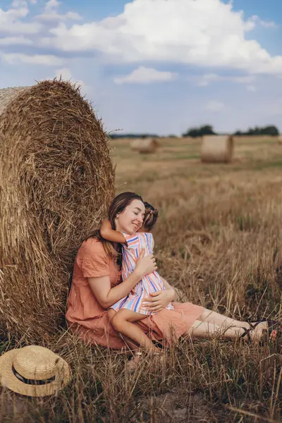 Mother and daughter sitting next to a bale of straw on a harvested wheat field. against the background of other bales. Daughter pressed against the mothers face