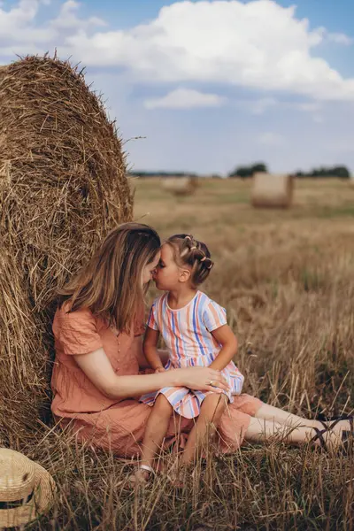 Mother and daughter sitting near a bale of straw on a harvested wheat field. against the background of other bales. Daughter kisses mom in the nose