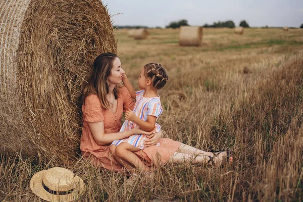 Mother and daughter sitting near a bale of straw on a harvested wheat field. against the background of other bales. Daughter touches mom by the face