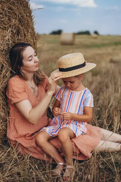Genç anne ve kız hasat edilen buğday tarlasında saman balyasının yanında oturuyorlar. Diğer balyaların arka planına karşı. Annem kızına saman anahtarı takıyor.