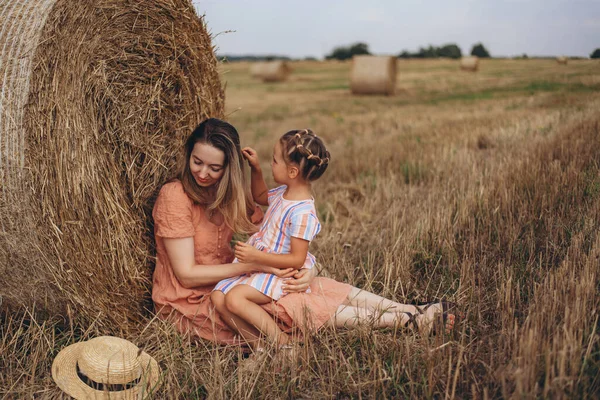 Mother and daughter sitting near a bale of straw on a harvested wheat field. against the background of other bales. Daughter touches mom by her hair