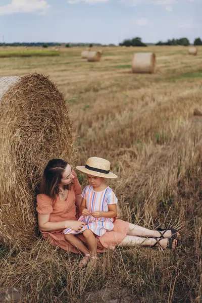 Genç anne ve kız hasat edilen buğday tarlasında saman balyasının yanında oturuyorlar. Diğer balyaların arka planına karşı. Kızım başka tarafa bakıyor.