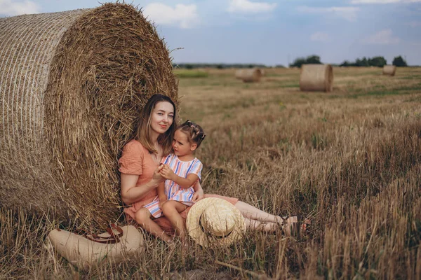 Genç anne ve kız hasat edilen buğday tarlasında saman balyasının yanında oturuyorlar. Kızı annesinin elini tutuyor..