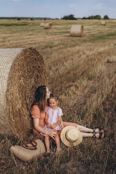 Genç anne ve kız hasat edilen buğday tarlasında saman balyasının yanında oturuyorlar. Yanlarında hasır bir çanta ve bir şapka var.