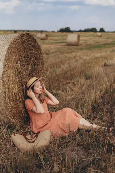 Uzun elbiseli ve uzun saçlı güzel bir kız sevimli gülüyor hasat edilmiş buğday tarlasında saman balyasında oturuyor. Şapkayı tutan kız.