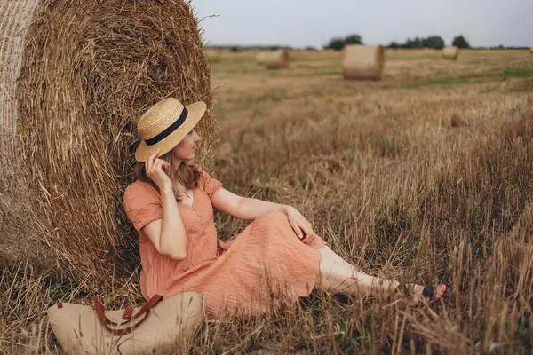 Uzun elbiseli ve uzun saçlı güzel bir kız sevimli gülüyor hasat edilmiş buğday tarlasında saman balyasında oturuyor. Kız hasır şapka için elini hareket ettiriyor.