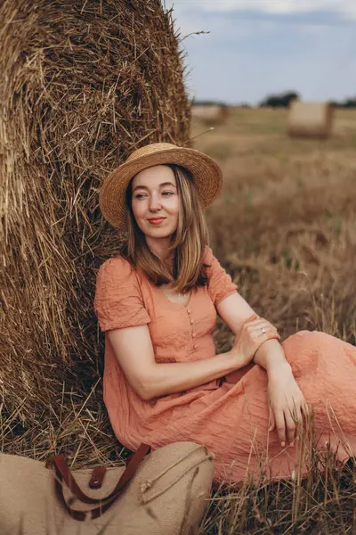 Uzun elbiseli, uzun saçlı, tatlı sırıtan güzel bir kız bir saman balyası üzerinde oturuyor. Kız başka yere bakar ve bir şeyler düşünür.