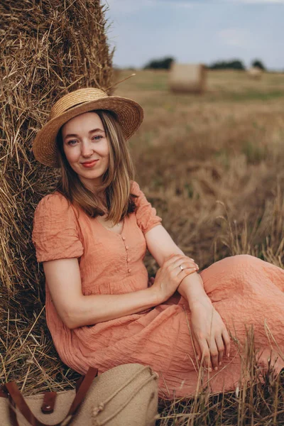Uzun elbiseli, uzun saçlı, tatlı sırıtan güzel bir kız bir saman balyası üzerinde oturuyor. Kız kameraya bakıyor.