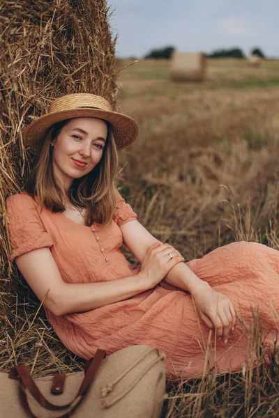 Uzun elbiseli, uzun saçlı, tatlı sırıtan güzel bir kız bir saman balyası üzerinde oturuyor. Kız kameraya bakıyor.