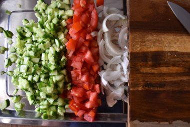 Üç sıra doğranmış sebze: salatalıklar, domatesler ve soğanlar demir tepside, tahta üst görünümün yanında. 