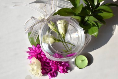 Yuvarlak bir vazoda beyaz çiçekler farklı çiçeklerin arasında fiyonk ve beyaz arka planda mumlarla süslenmiş.