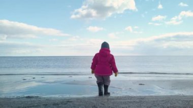 Küçük bir kız baharın başında sahilde yürüyor ve sığ sularda mutlu bir şekilde zıplıyor. Güneşli, güzel bir bahar günü, biraz bulutlu..